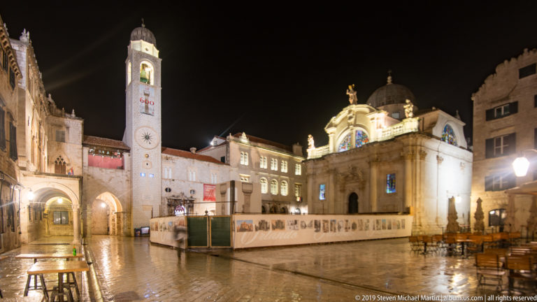 Dubrovnik City Bell Tower beside the illuminated Church of St. Blaise across wet Luža Square at night.