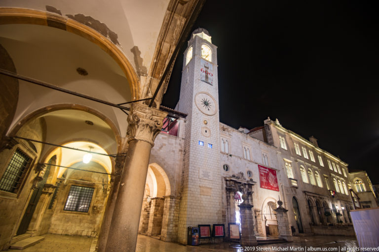 Dubrovnik City Bell Tower seen beyond a stone arcade, illuminated façades, and wet paving at night.