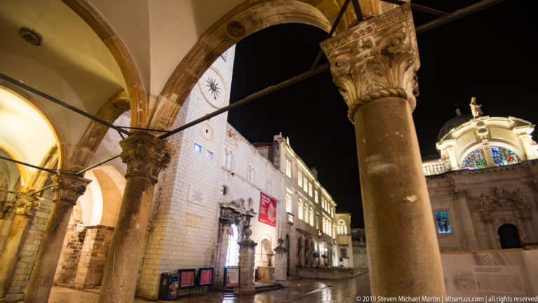 Carved arcade column and vault framing Dubrovnik’s City Bell Tower and illuminated square at night.