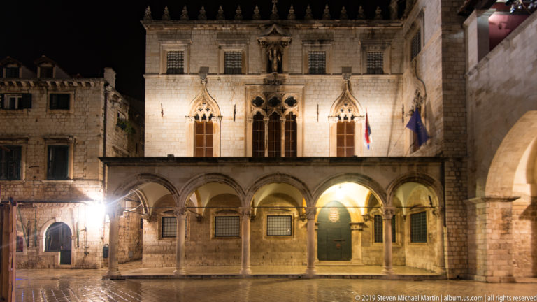 Illuminated Sponza Palace façade with ground-floor arcade, Gothic windows, central statue, and flags at night.