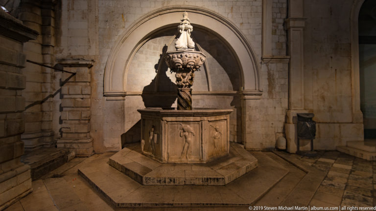 Small Onofrio Fountain with sculpted upper element and polygonal basin set beneath a stone arch at night.