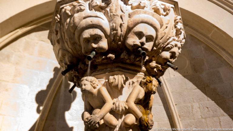Close-up of a carved grotesque mask, water spout, putti, and foliage on Dubrovnik’s Small Onofrio Fountain.