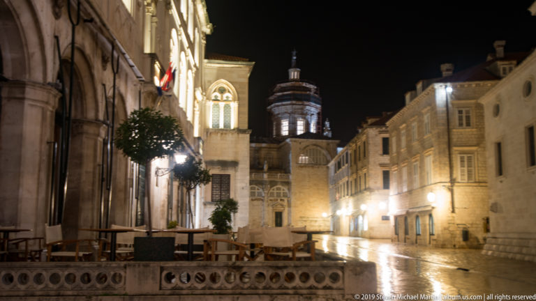 Quiet wet Luža Square lined by illuminated historic buildings with Dubrovnik’s City Bell Tower at the far end.