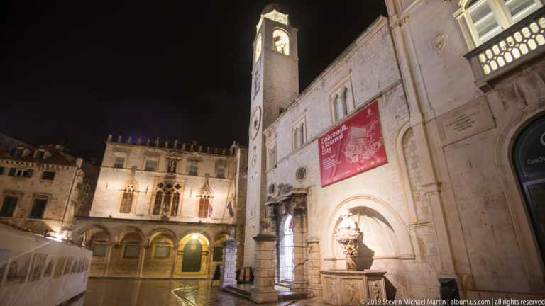Dubrovnik City Bell Tower rising above illuminated stone façades, banners, and arched entrances at night.