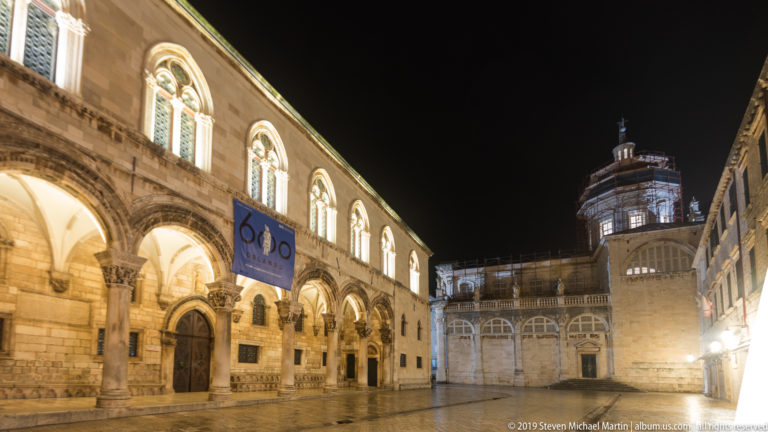 Rector’s Palace arcade receding toward Dubrovnik’s illuminated City Bell Tower across wet paving at night.