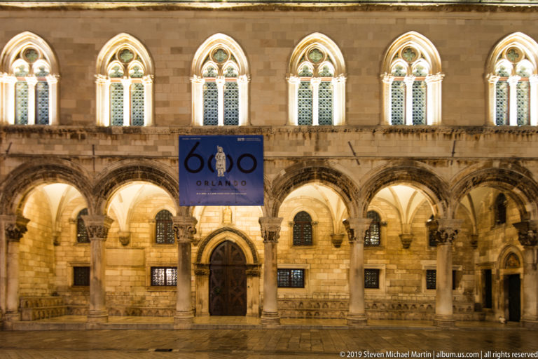 Illuminated Rector’s Palace façade with ground-floor arcade, Gothic upper windows, central doorway, and wet stone pavement.