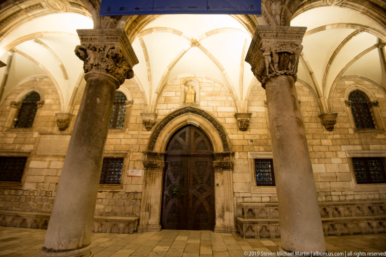 Arched wooden doorway framed by columns, carved capitals, and ribbed vaults in the Rector’s Palace loggia.