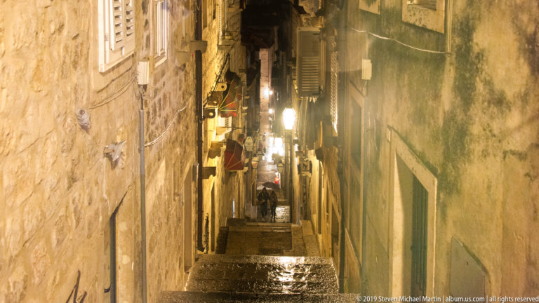 Steep wet stone steps rising between narrow historic buildings with lamps, shutters, signs, and weathered walls at night.