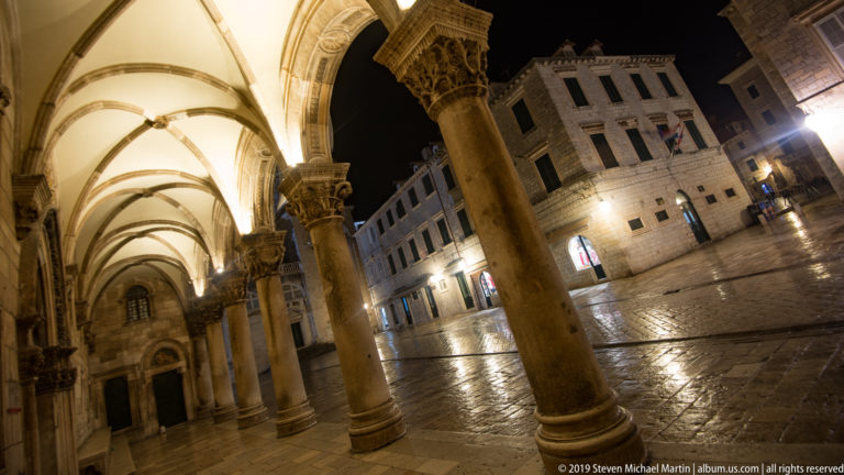 Stone columns and pointed vaults of the Rector’s Palace loggia opening onto a wet illuminated square at night.