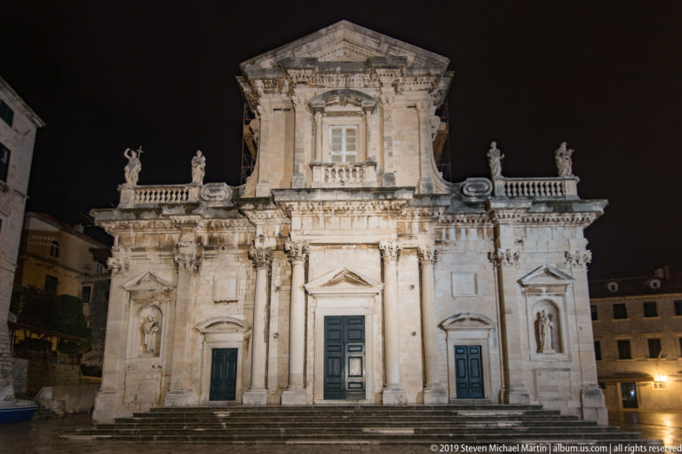 Illuminated Baroque façade of Dubrovnik Cathedral with three portals, statues, columns, and a central pediment at night.