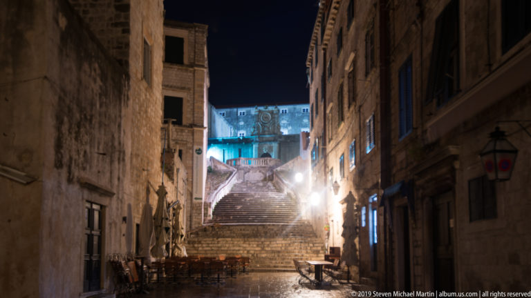 Jesuit Stairs rising between stone buildings toward the illuminated upper Old City, with empty café tables below.