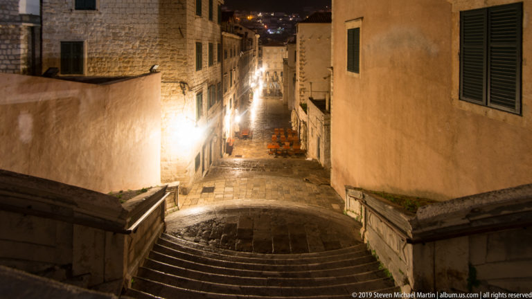 Broad wet stone stairway descending between tall historic buildings into a lamp-lit Dubrovnik lane at night.