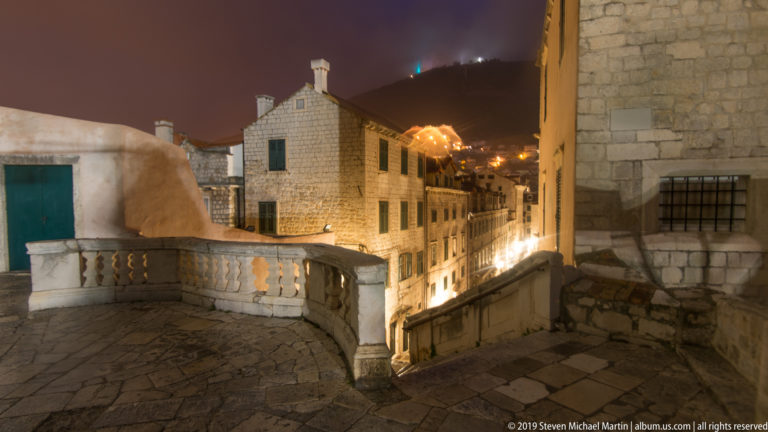 Stone terrace and balustrade overlooking a descending lamp-lit lane and historic masonry houses in Dubrovnik at night.