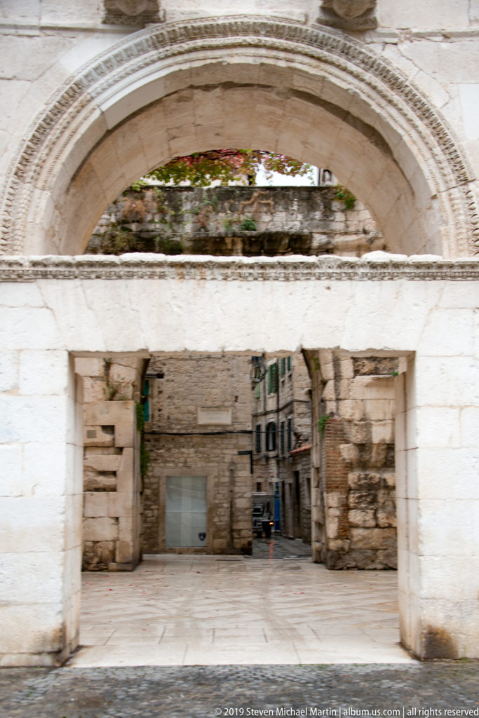 View beneath a large carved stone arch toward a paved courtyard and narrow old-town street beyond.