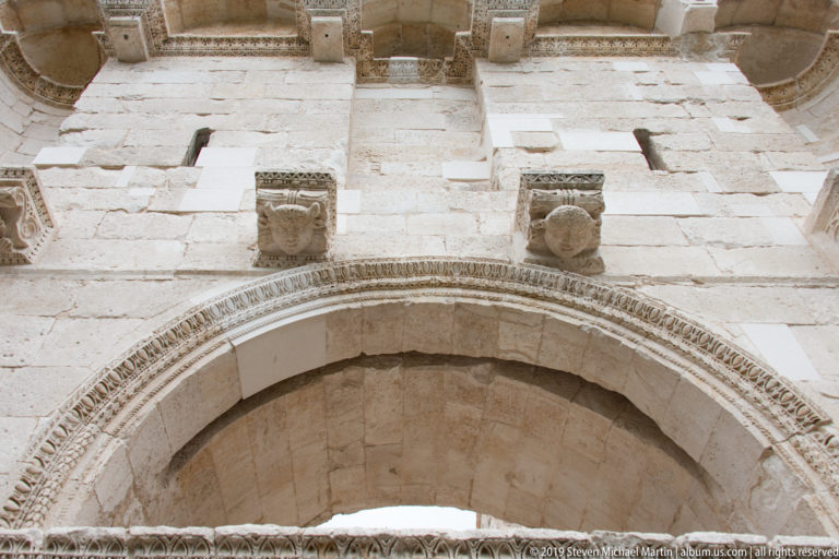 Large semicircular stone arch with patterned moldings and two carved human heads above it.