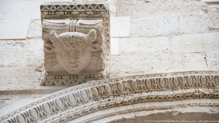 Close view of a carved human head above patterned stone moldings on an arched portal.