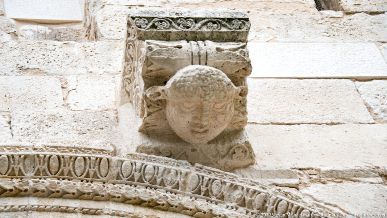 Weathered carved human head projecting above bands of geometric ornament on a pale stone arch.