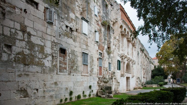 Long weathered stone wall with blocked openings, arched windows, doors and landscaped strip along its base.