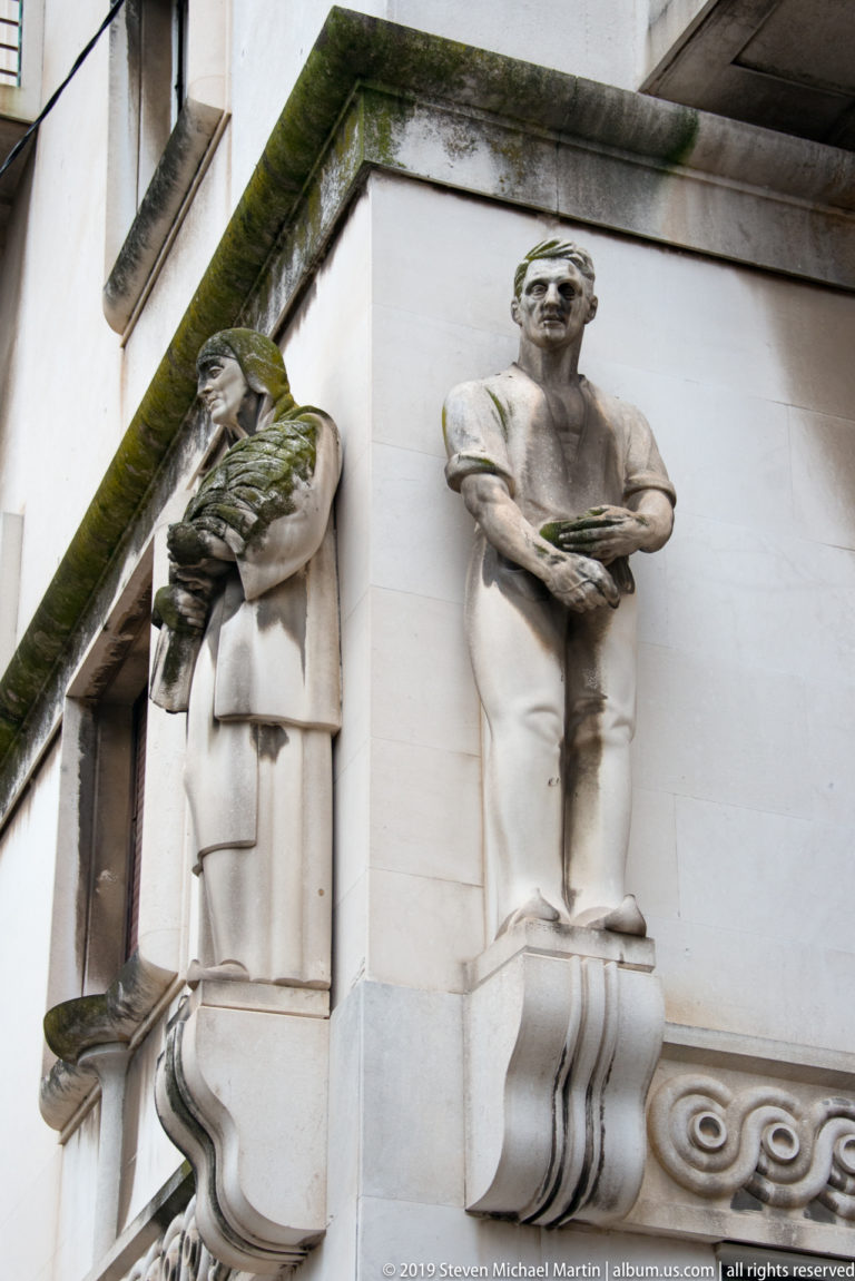 Two weathered stone human figures attached to a pale façade, one in a niche and one projecting on a bracket.