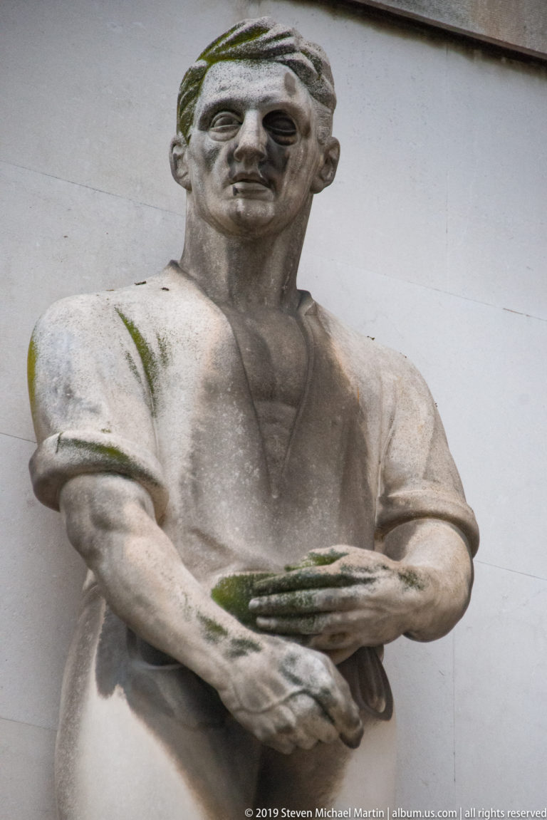 Weathered stone male figure holding a shallow rectangular object against a pale wall, with moss on the carving.