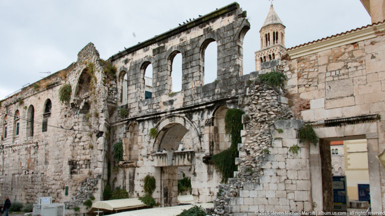 Ruined stone wall with tall arched openings and vegetation, with the Cathedral of Saint Domnius bell tower behind.