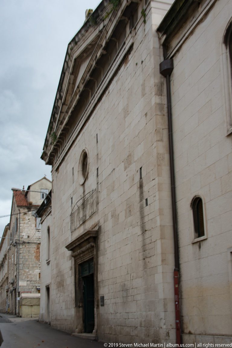 Plain pale-stone church façade with small circular window and dark rectangular doorway on a narrow street.