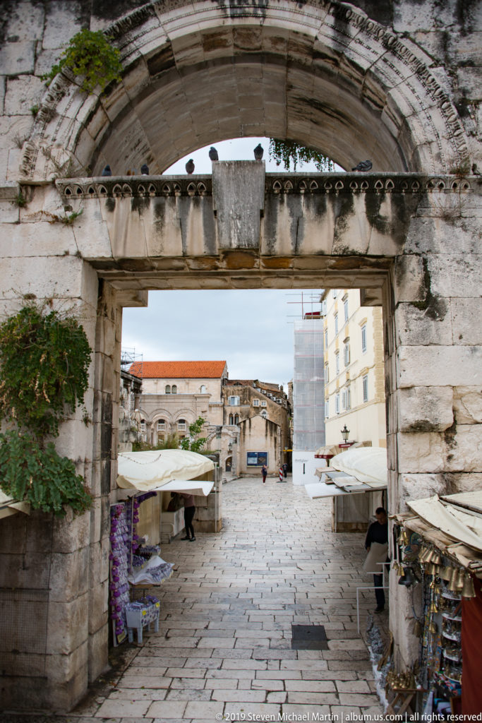 Large stone arch framing a paved pedestrian passage with market stalls and buildings beyond.