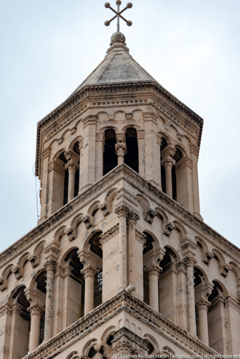 Upper levels of a pale stone bell tower with repeated arcades, paired columns, roof and cross.
