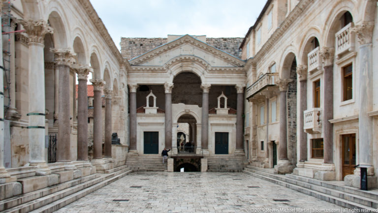 Stone-paved Peristyle court lined with colonnades and arcaded façades leading toward monumental architecture.
