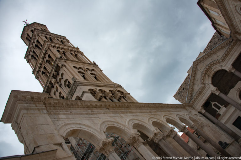 Upward view through pale stone arches toward the Cathedral of Saint Domnius bell tower under a cloudy sky.
