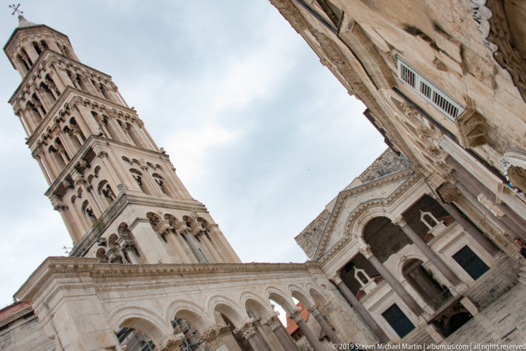 Low-angle view of the Cathedral of Saint Domnius bell tower rising above arcaded pale stone architecture.