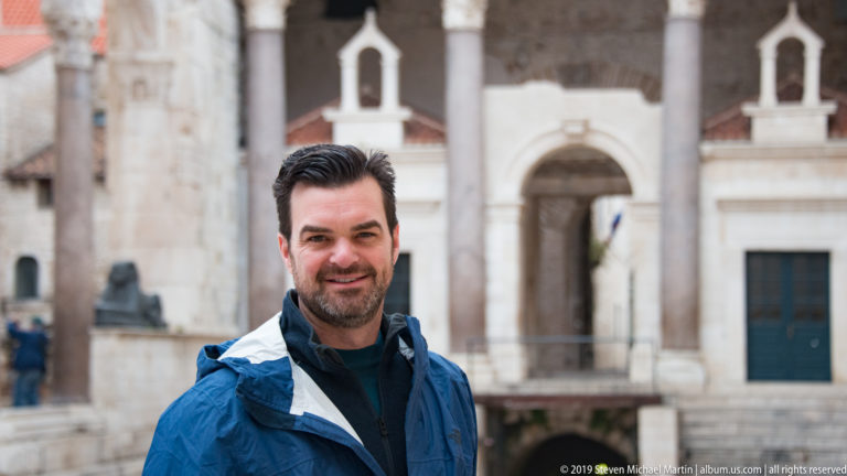 Smiling man in a blue jacket standing before pale stone arcades and a monumental doorway in Split.