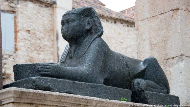Side view of a dark stone sphinx with extended forepaws and human head against pale limestone walls.