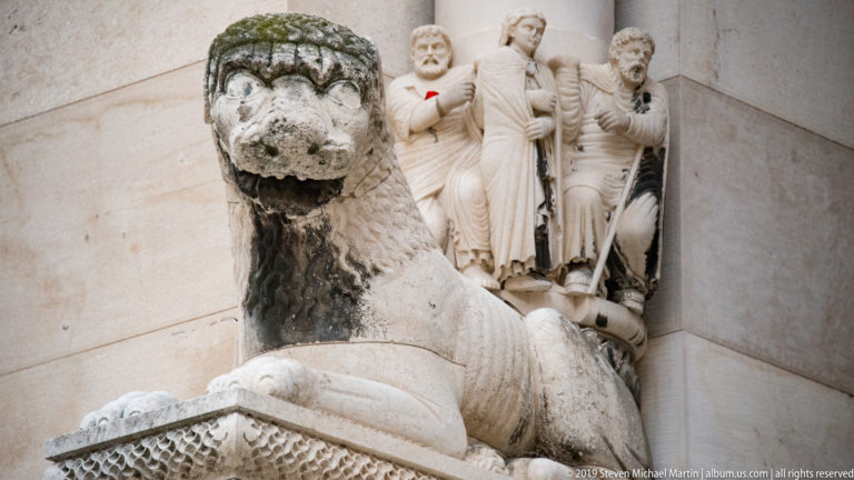 Weathered stone sphinx with human head and lion body in front of carved architectural figures at Diocletian’s Palace.