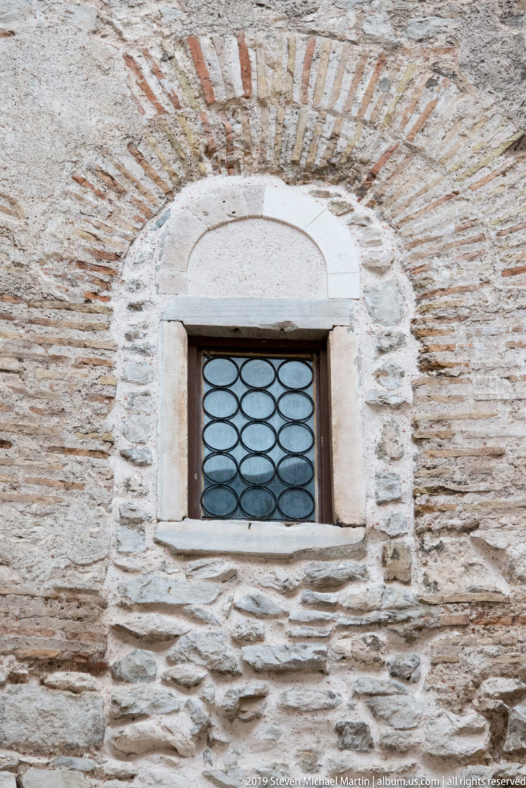Small round-arched window with pale stone surround and metal lattice set into weathered stone-and-brick masonry.