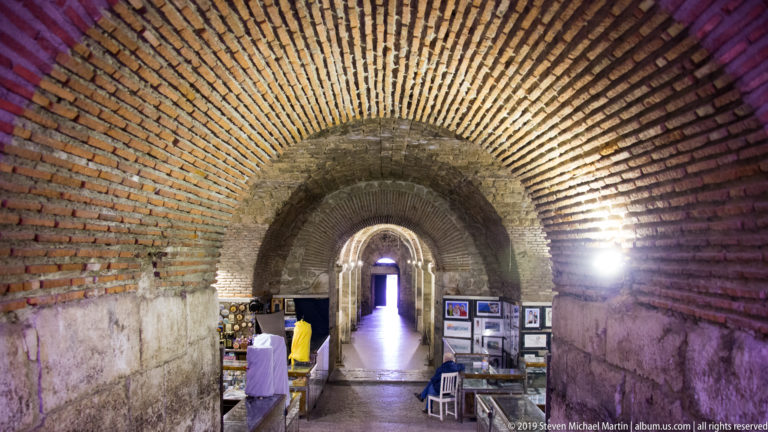 Brick barrel-vaulted passage with successive arches, displays, and a bright exit beneath Diocletian’s Palace.