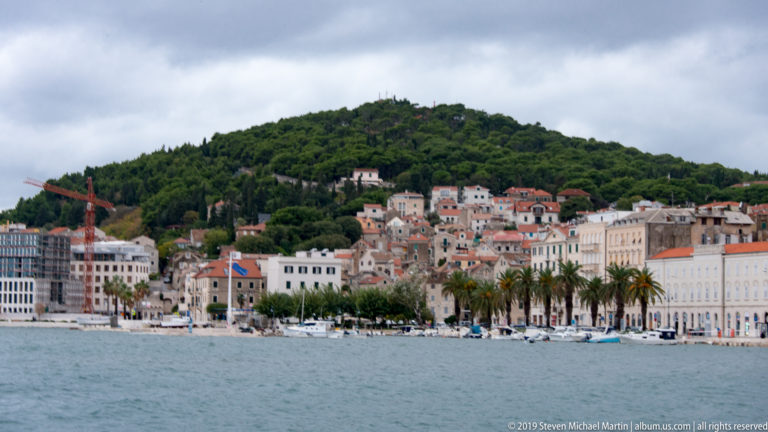 Harbor panorama of Split’s palm-lined waterfront, pale buildings, hillside houses, and moored boats.