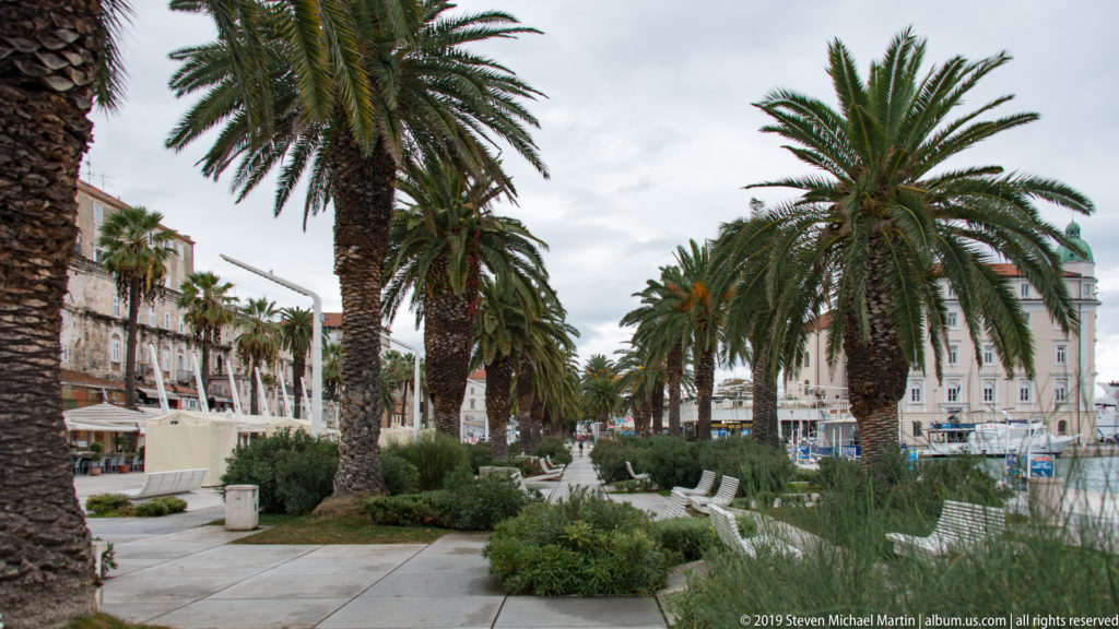 Palm-lined Split waterfront promenade with benches, planted beds, walkway, and marina at right.