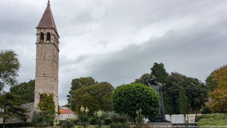 Tall square stone bell tower rising above trees, with the Gregory of Nin statue visible nearby.