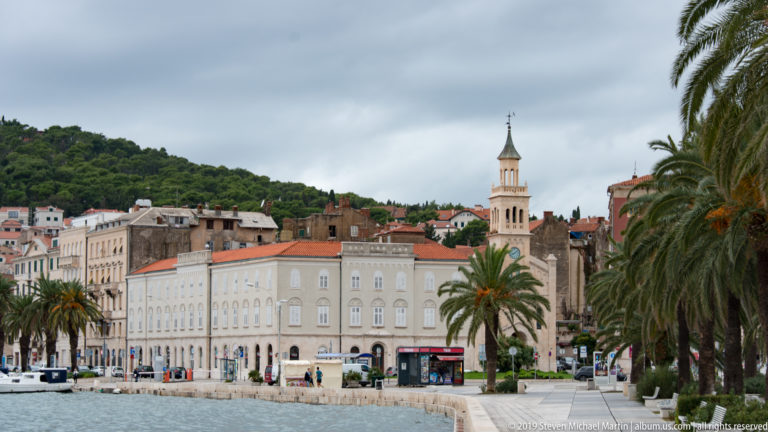 Split waterfront promenade with palms, pale buildings, buses, and a bell tower beneath an overcast sky.