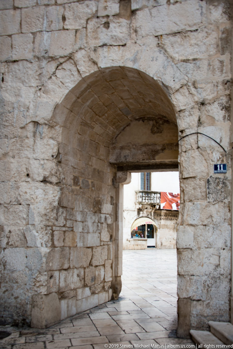 Shadowed low stone archway opening through thick block masonry toward a bright paved square.