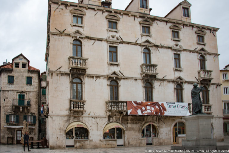 Tall pale-stone façade with small balconies, arched shopfronts, and a contemporary exhibition banner.