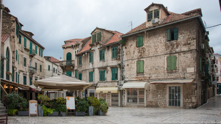 Weathered stone houses with green shutters, balconies, red tile roofs, and restaurant seating at street level.