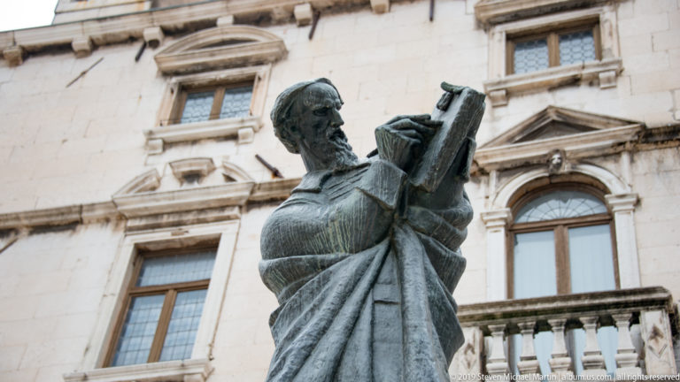 Low-angle view of a bronze robed man holding an open book or tablet before a historic façade.