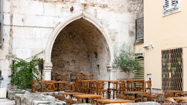 Pointed Gothic stone recess filled with stacked wooden café chairs beside a modern building wall.