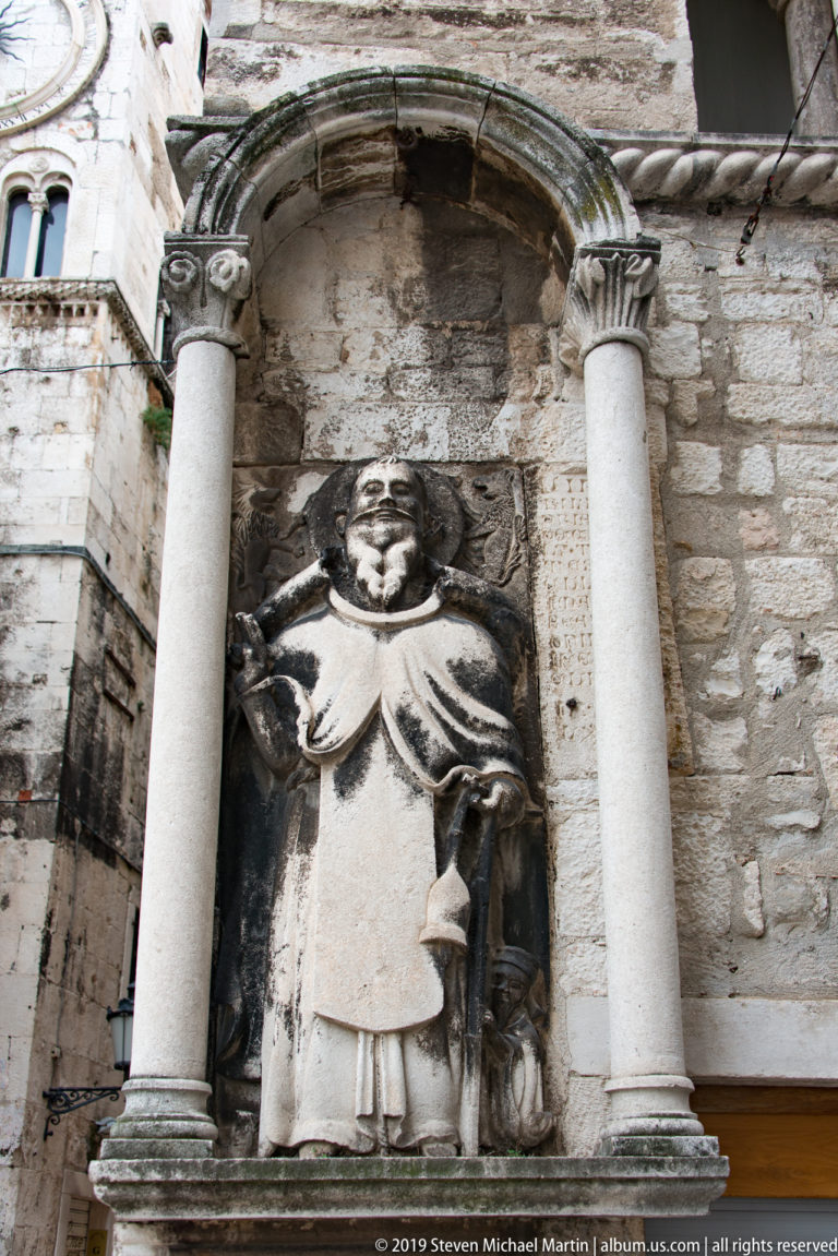 Weathered robed stone figure standing in a shallow niche between two pale columns.