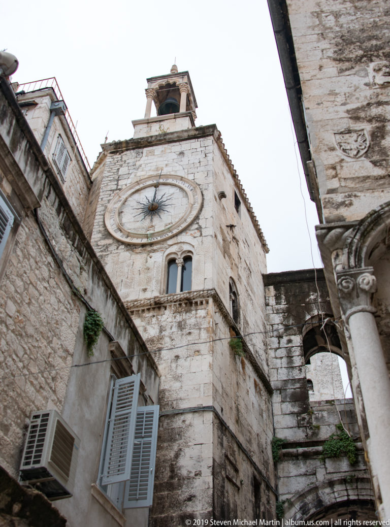 Upward view of a weathered stone clock tower framed tightly by neighboring historic buildings.