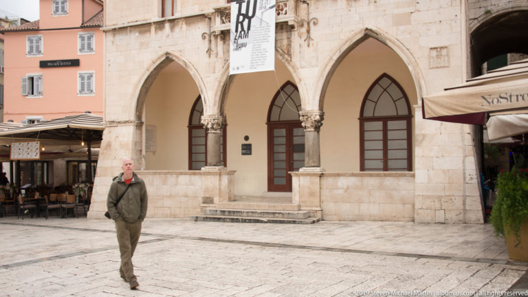 Man walking before two tall pointed stone arches with slender columns on a historic façade.