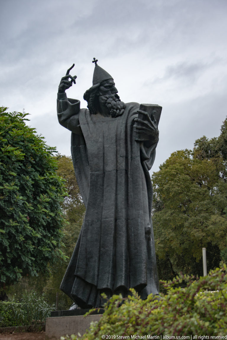 Large dark bronze statue of Bishop Gregory of Nin raising one hand and holding a book amid trees.