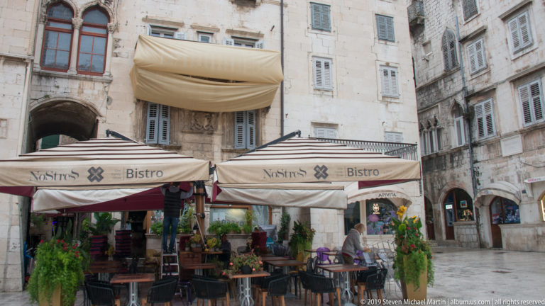 Outdoor café with striped awnings, plants, and tables beneath pale stone façades and shuttered windows.